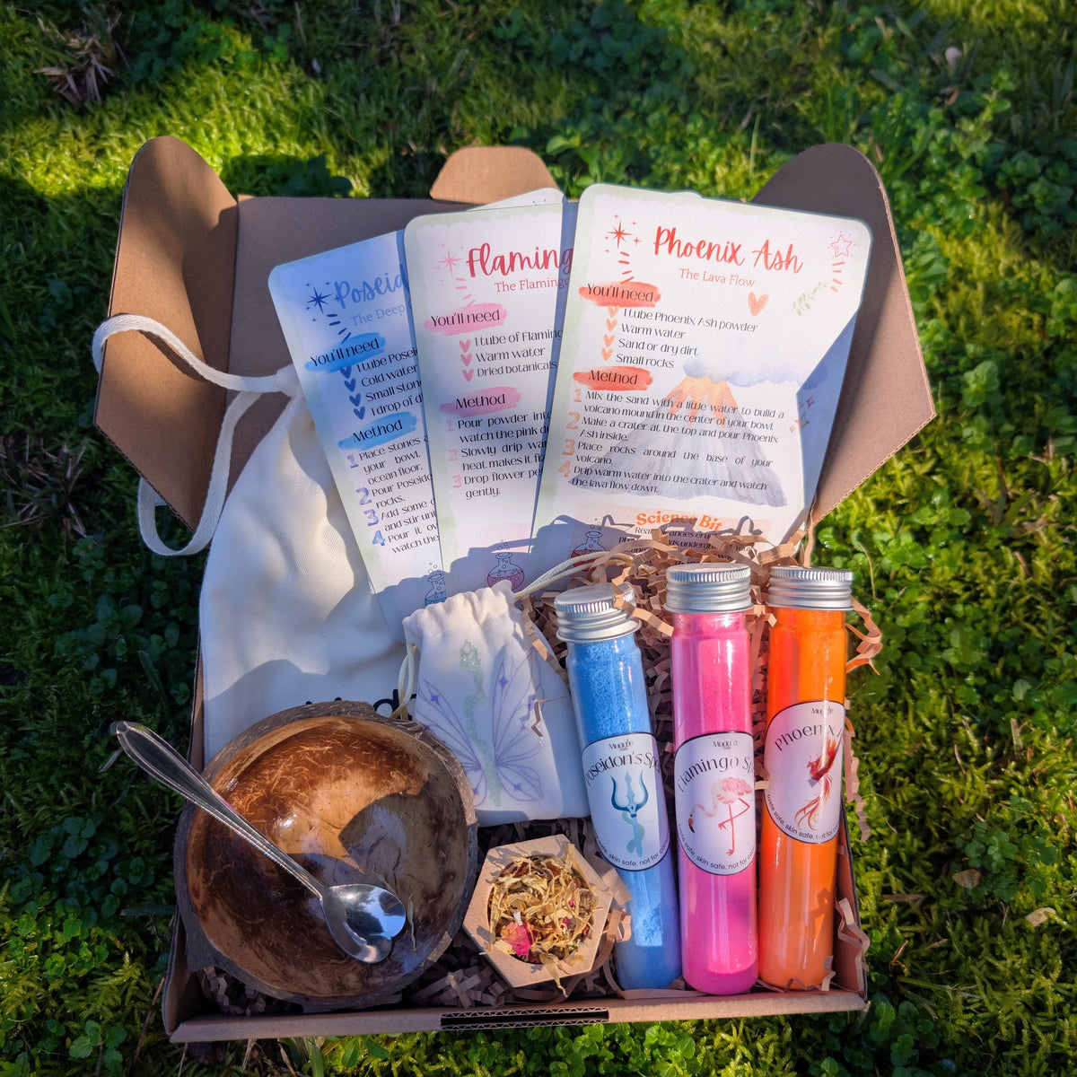 Open Muddle & Play kit box with story cards, test tubes, a coconut bowl and a drawstring bag, on grass