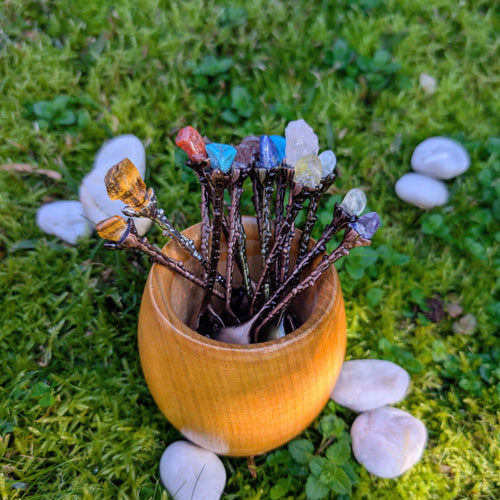 Gemstone-topped spoons standing in a wooden cup on grass among white stones