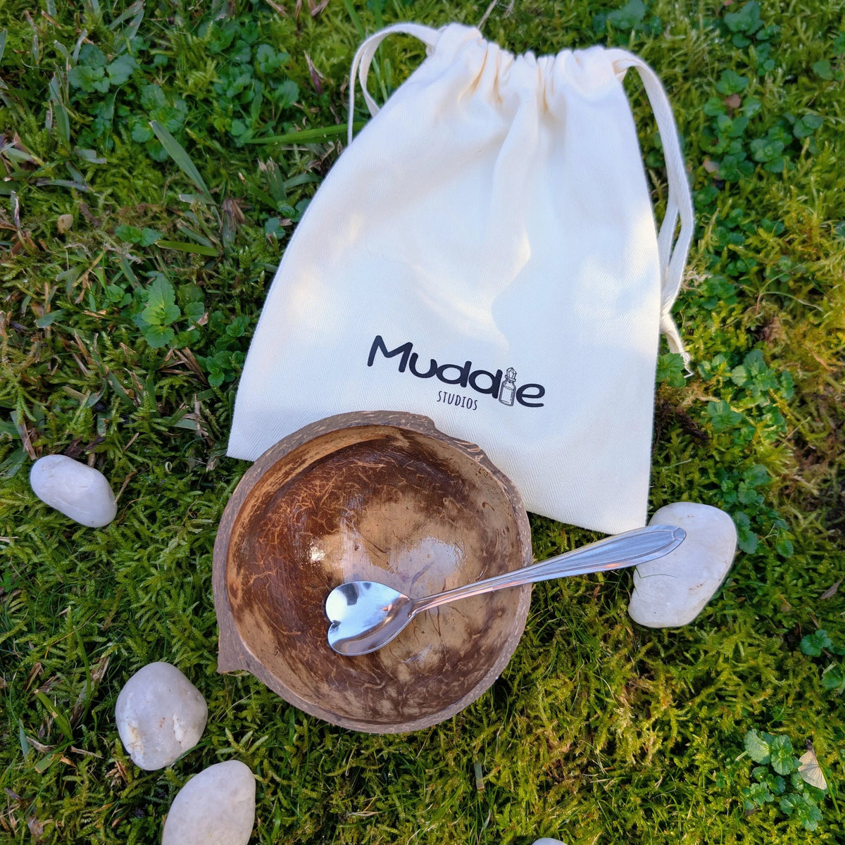 Coconut shell bowl with a heart-shaped teaspoon and a Muddle Studios drawstring bag, on grass