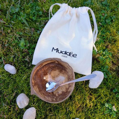 Coconut shell bowl with a heart-shaped teaspoon and a Muddle Studios drawstring bag, on grass