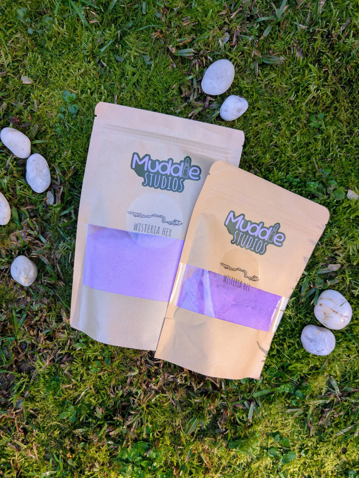 Two kraft pouches of Wisteria Hex purple fizzy powder on grass beside white stones
