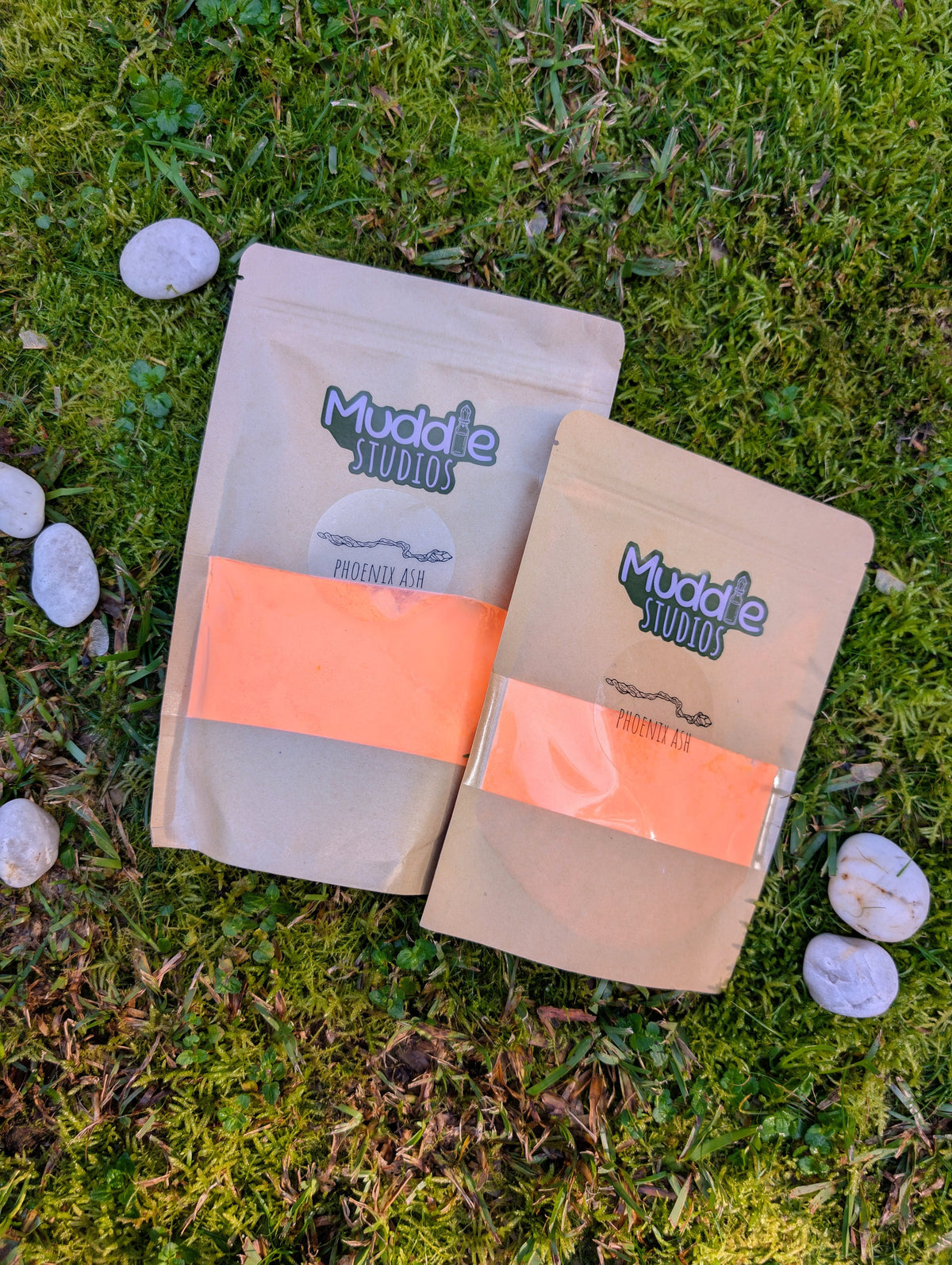Two kraft pouches of Phoenix Ash orange fizzy powder on grass beside white stones