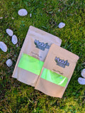 Two kraft pouches of Liquid Gold fizzy powder on grass beside white stones