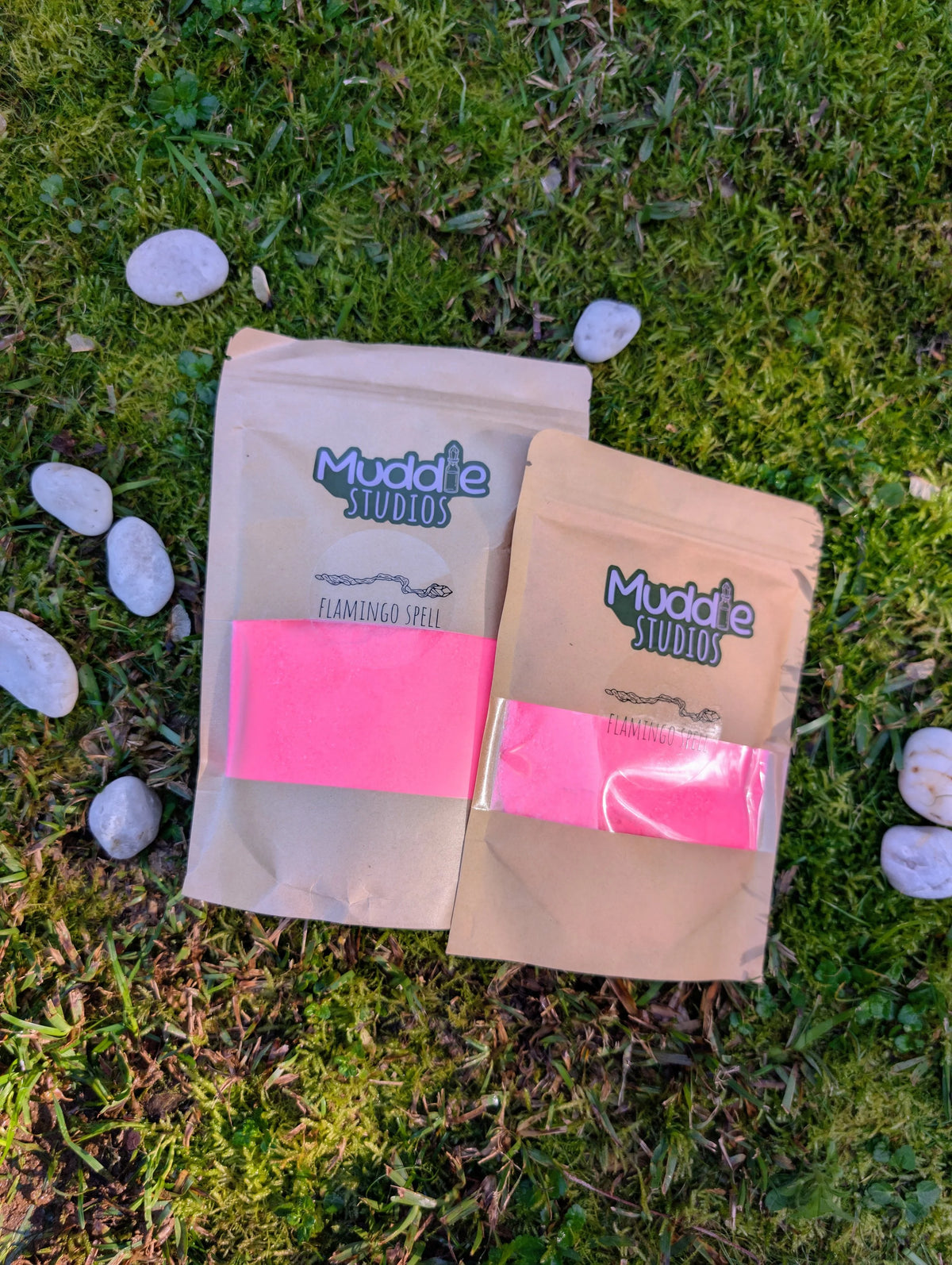 Two kraft pouches of Flamingo Spell pink fizzy powder on grass beside white stones