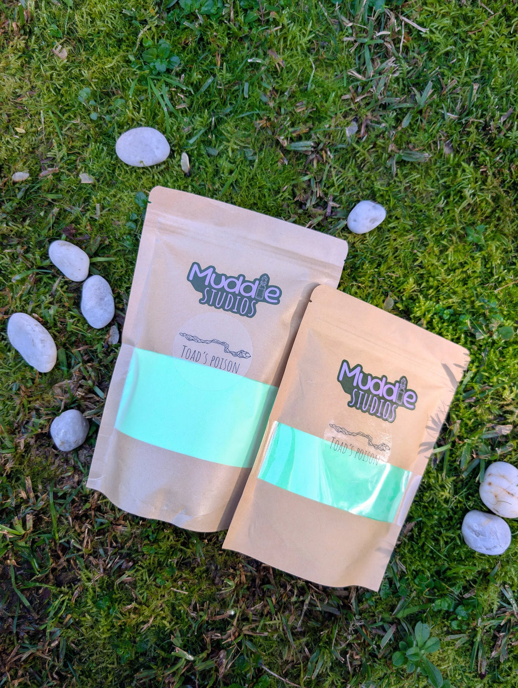 Two kraft pouches of Toad's Poison green fizzy powder on grass beside white stones 