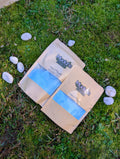 Two kraft pouches of Poseidon's Spell blue fizzy powder on grass beside white stones