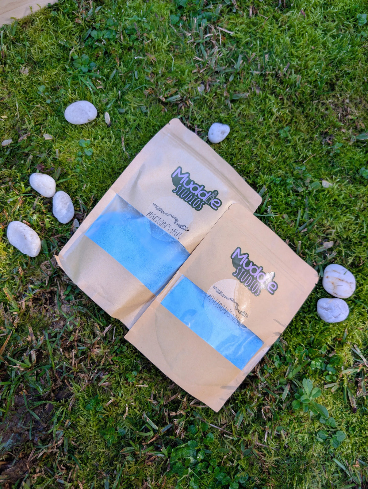Two kraft pouches of Poseidon's Spell blue fizzy powder on grass beside white stones
