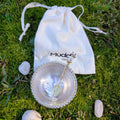 Stainless steel bowl with a spoon and a Muddle Studios drawstring bag, on moss and stones