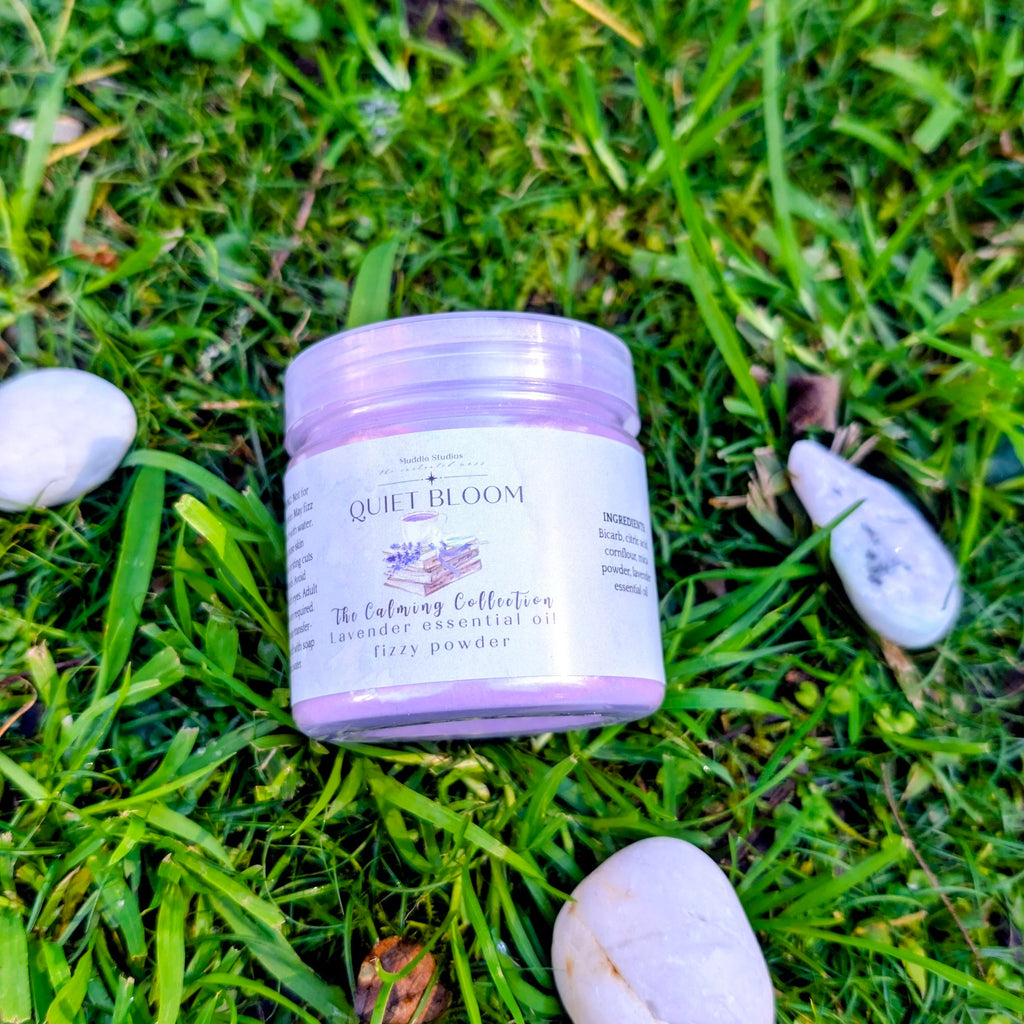 Quiet Bloom fizzy powder jar with a lavender label on grass beside white stones