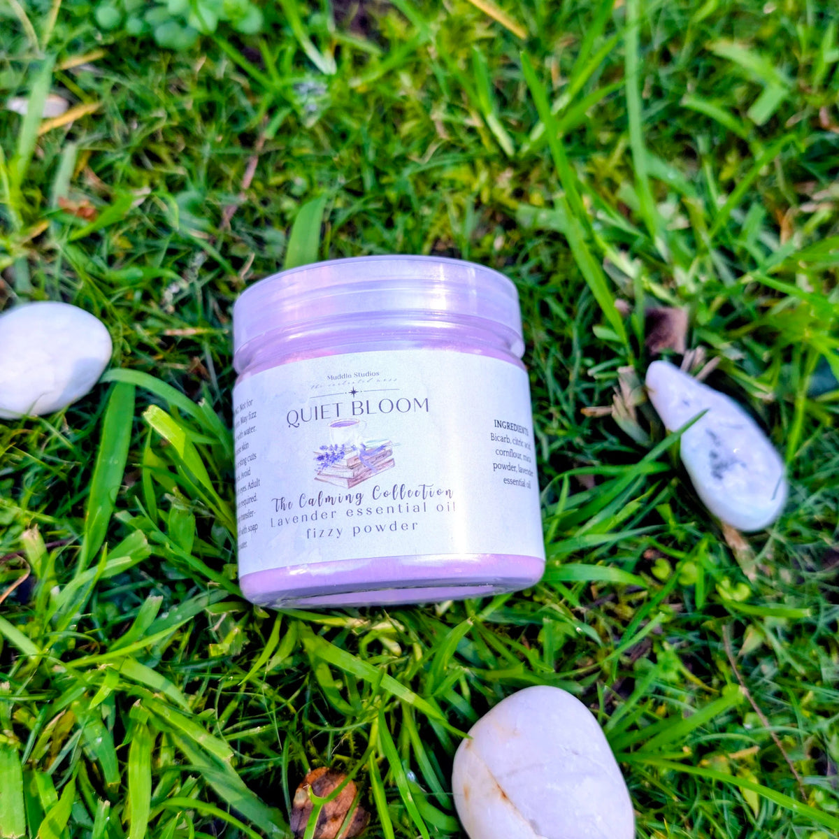 Quiet Bloom fizzy powder jar with a lavender label on grass beside white stones