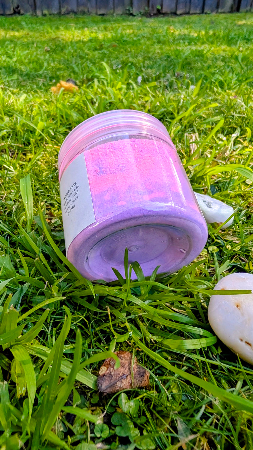 Open jar of Sunset Bloom, pink and purple fizzy powder, seen from above on grass 