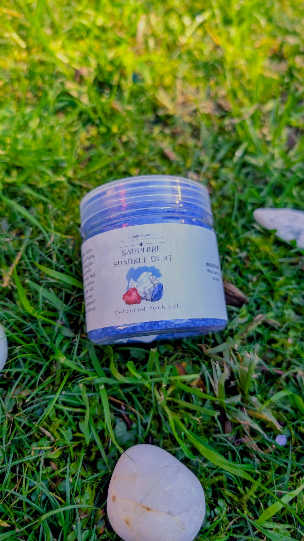 Sapphire Sparkle Dust jar with a blue label on grass beside a stone
