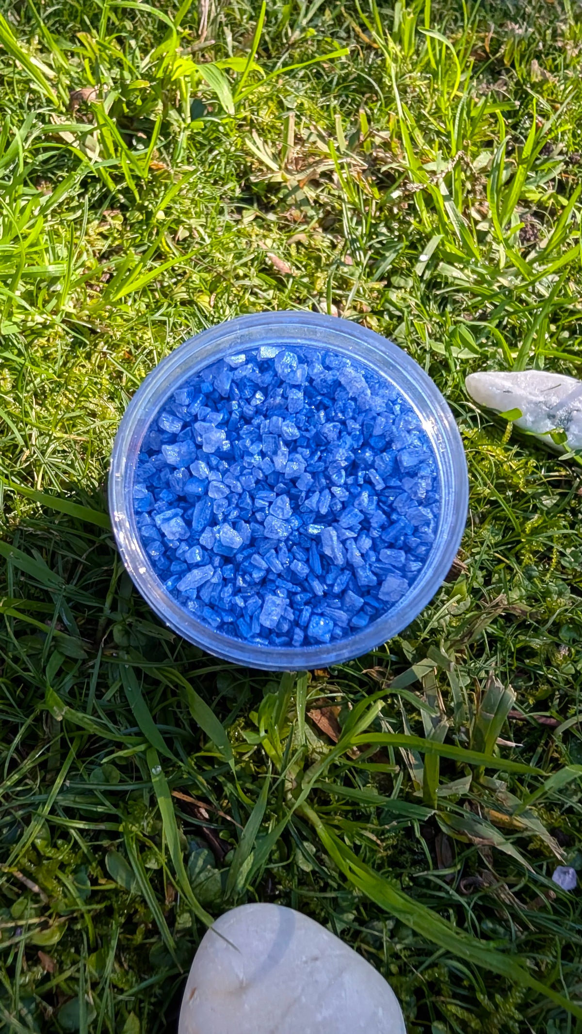 Open jar of Sapphire Sparkle Dust, blue mica-coated rock salt crystals, seen from above on grass