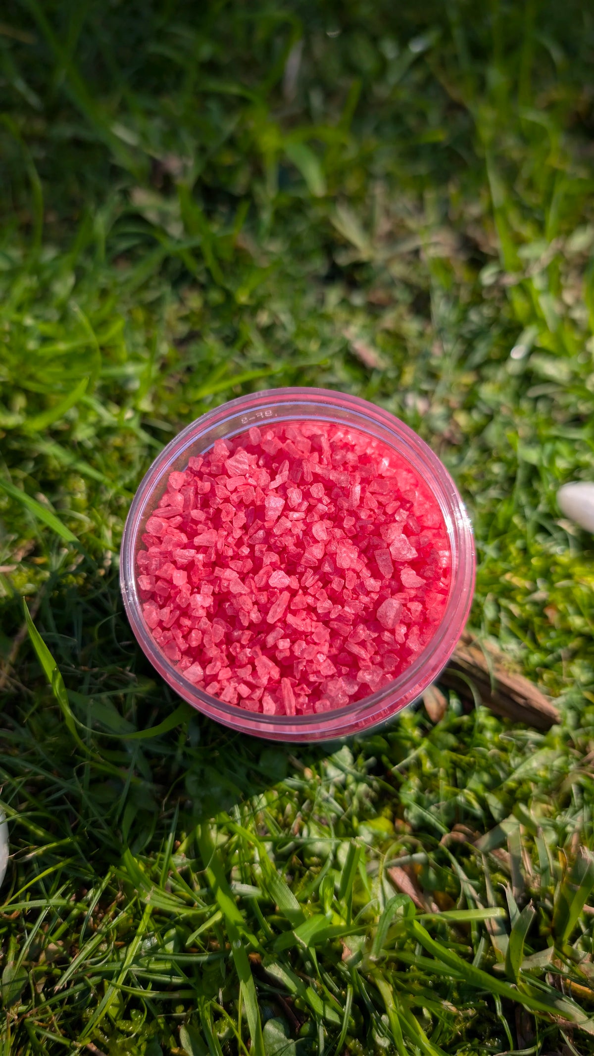 Open jar of Ruby Sparkle Dust, red mica-coated rock salt crystals, seen from above on grass