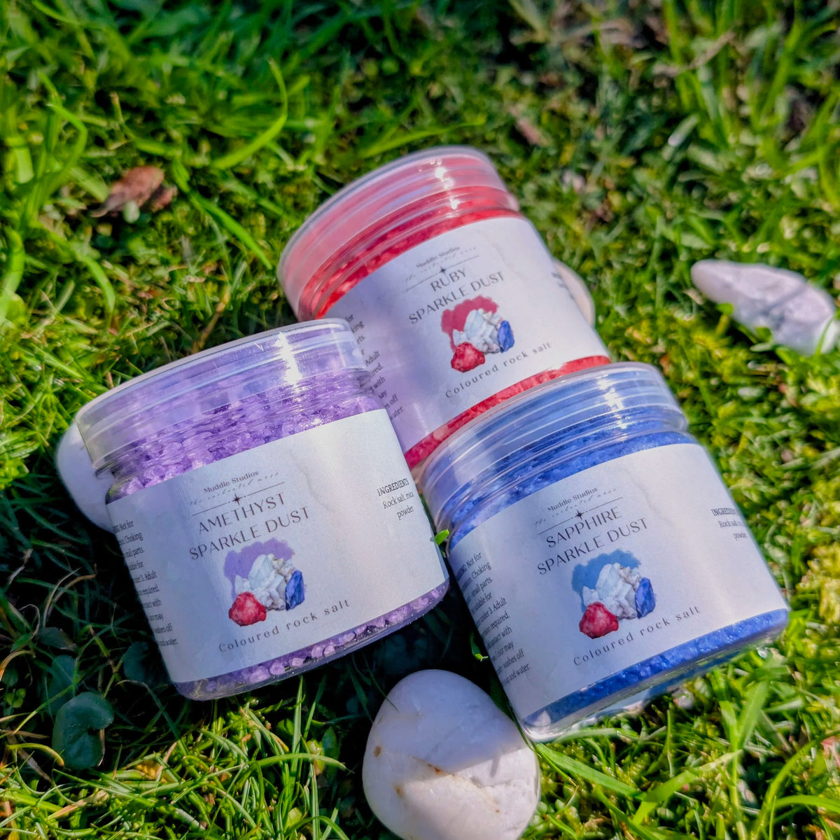 Sapphire, Amethyst and Ruby Sparkle Dust jars side by side on grass with a white stone