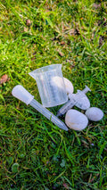 Clear plastic potion tool set with droppers, a spoon and a measuring cup on grass beside white stones