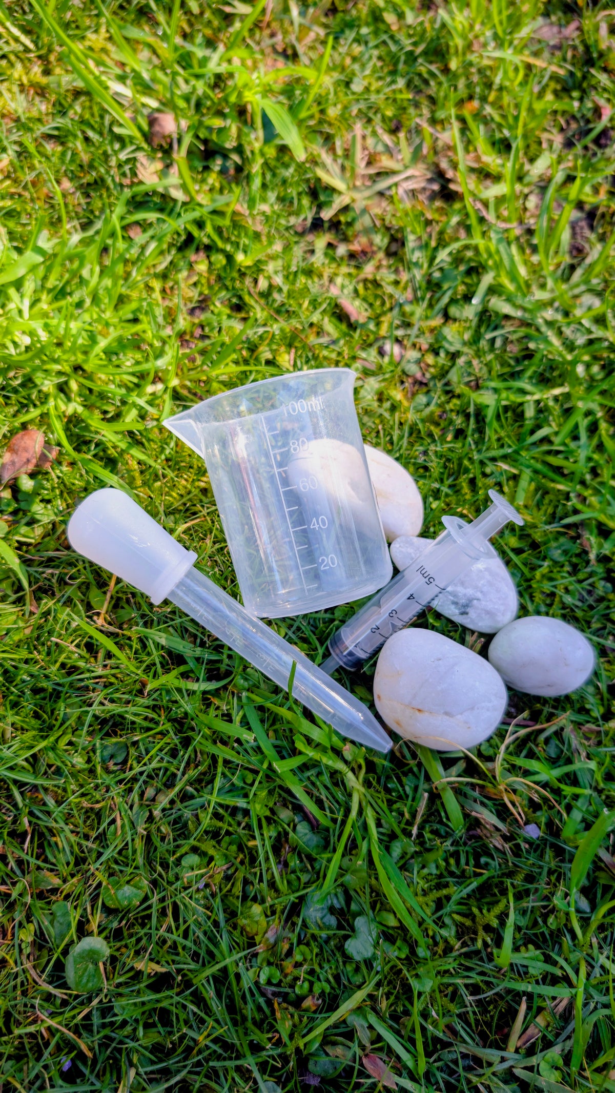 Clear plastic potion tool set with droppers, a spoon and a measuring cup on grass beside white stones