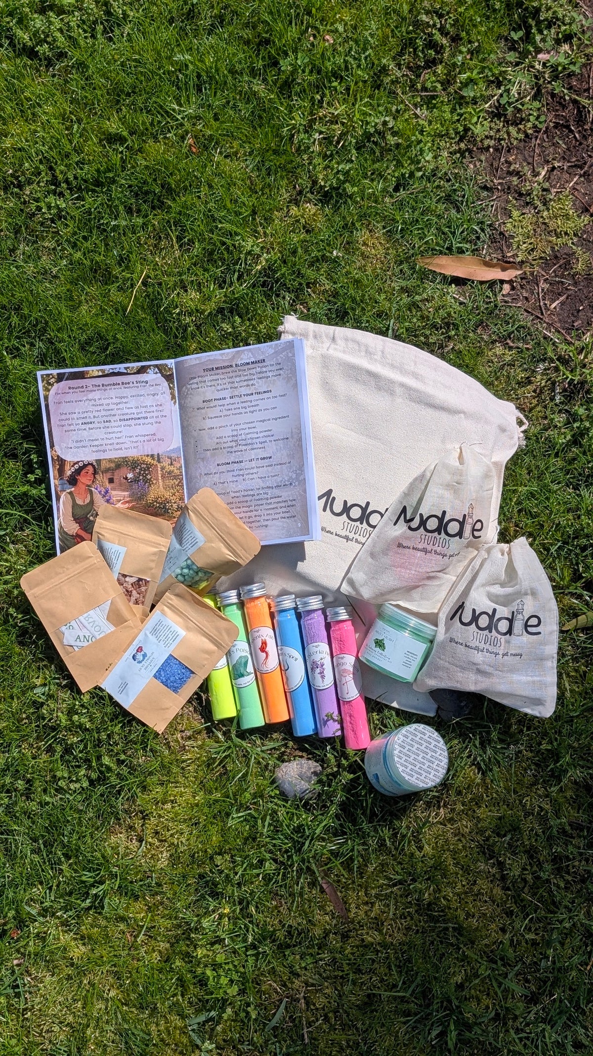 The Root & Bloom Kit laid out on grass with an open storybook, a drawstring Muddle bag, colourful test tubes, a jar and pouches