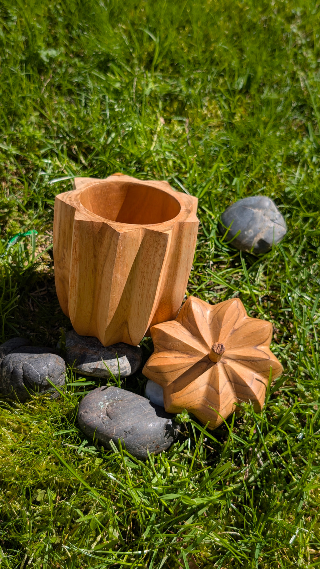 Nona wooden pot with its flower-shaped lid placed in front, on grass among stones