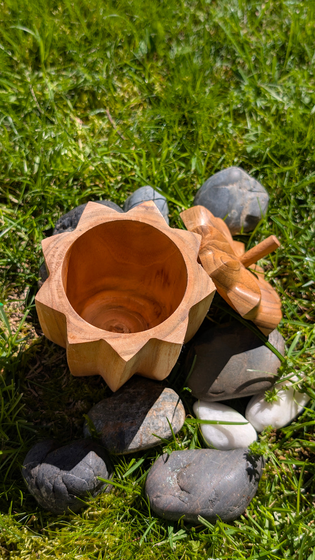 Nona wooden pot open from above with its flower-shaped lid beside it, on grass and stones