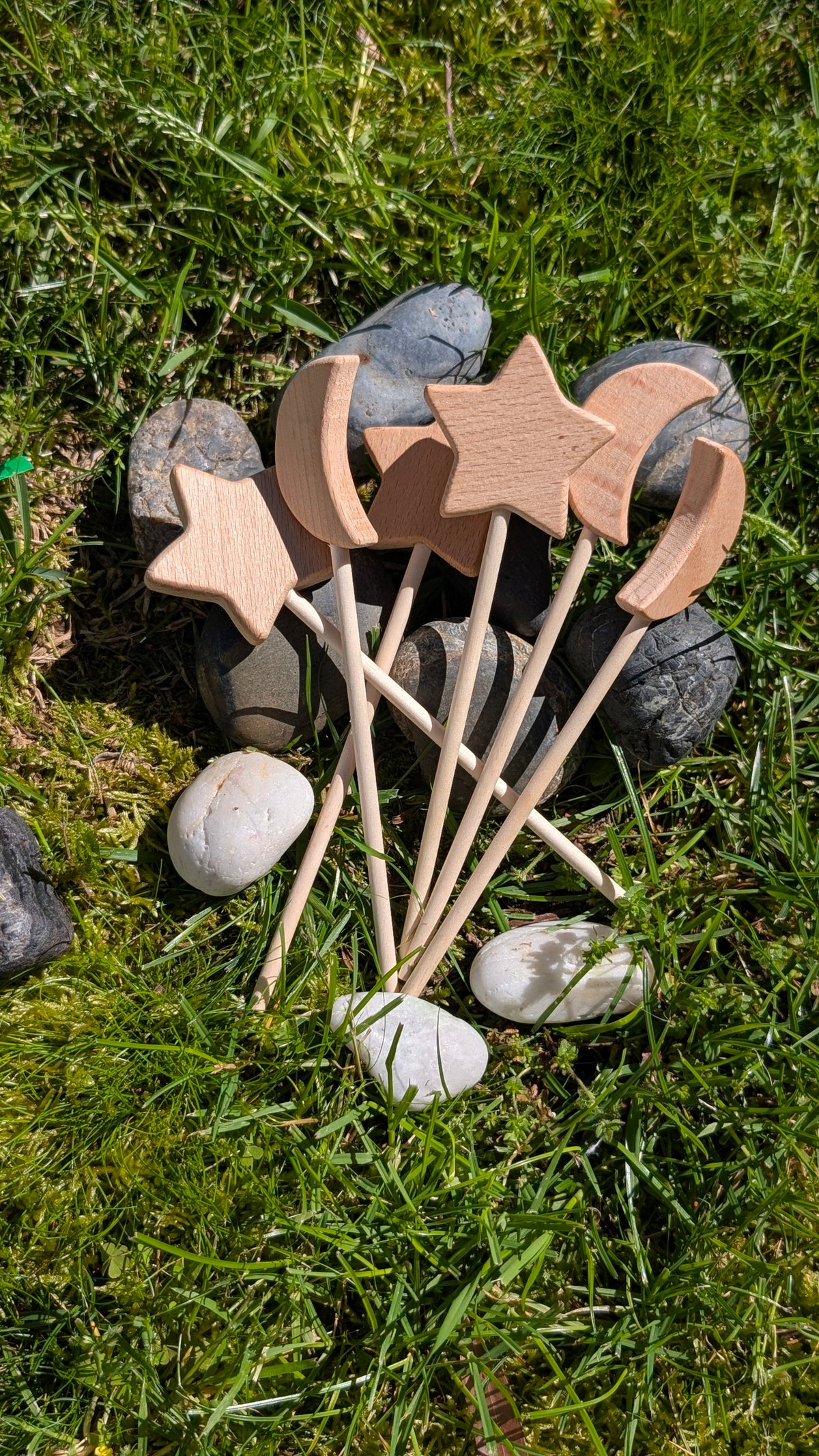 Five wooden fairy wands topped with a star, crescent moon and sun, laid on grass among stones